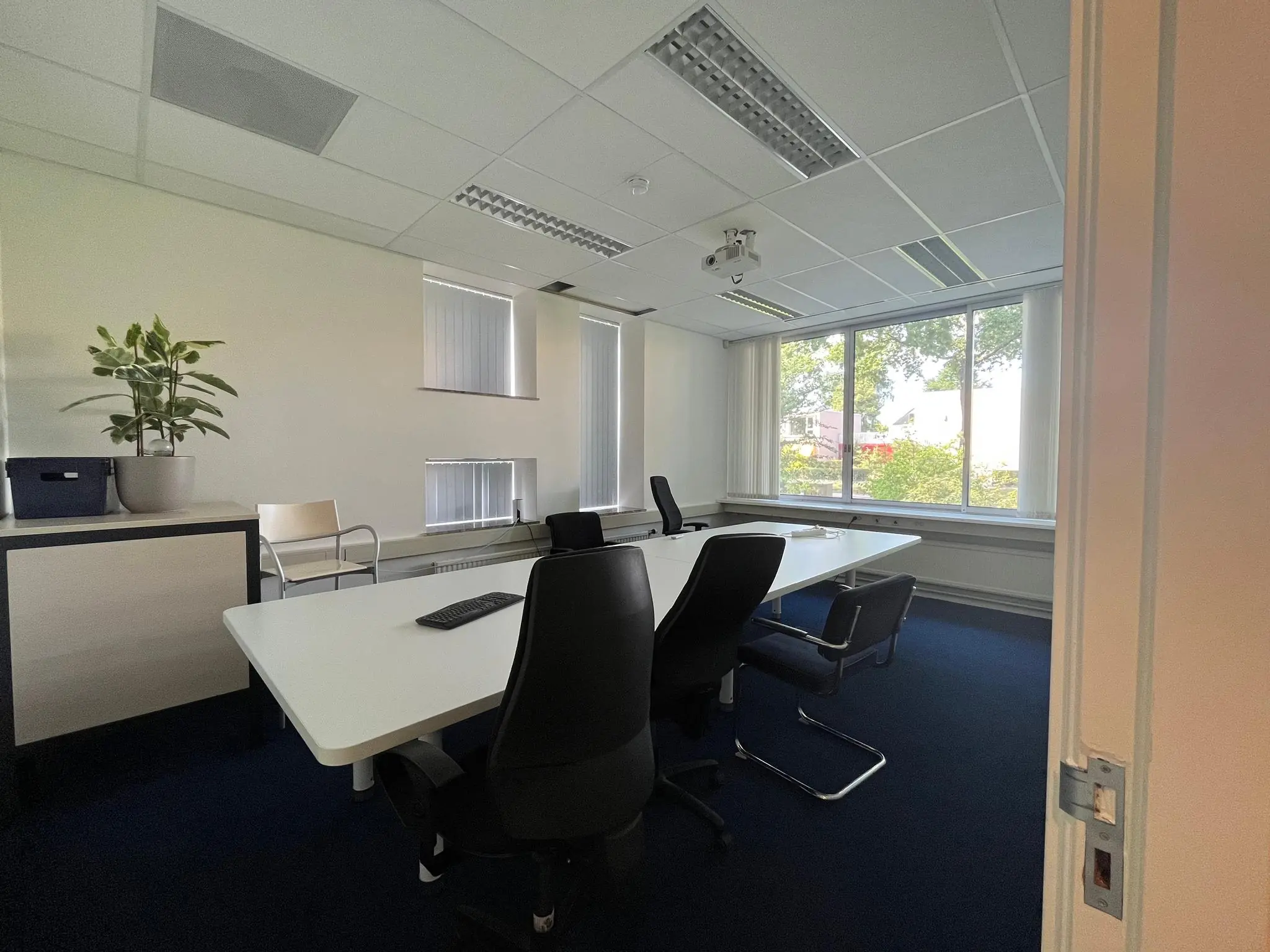 Vergaderruimte aan de Jan van Eycklaan met een lange witte tafel, zwarte stoelen en grote ramen met uitzicht op groen.